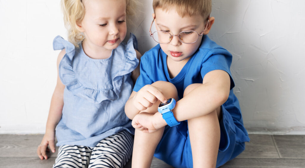 A little boy child in a blue T-shirt and glasses shows the girl his smart watch and presses a finger. Portrait against white wall.